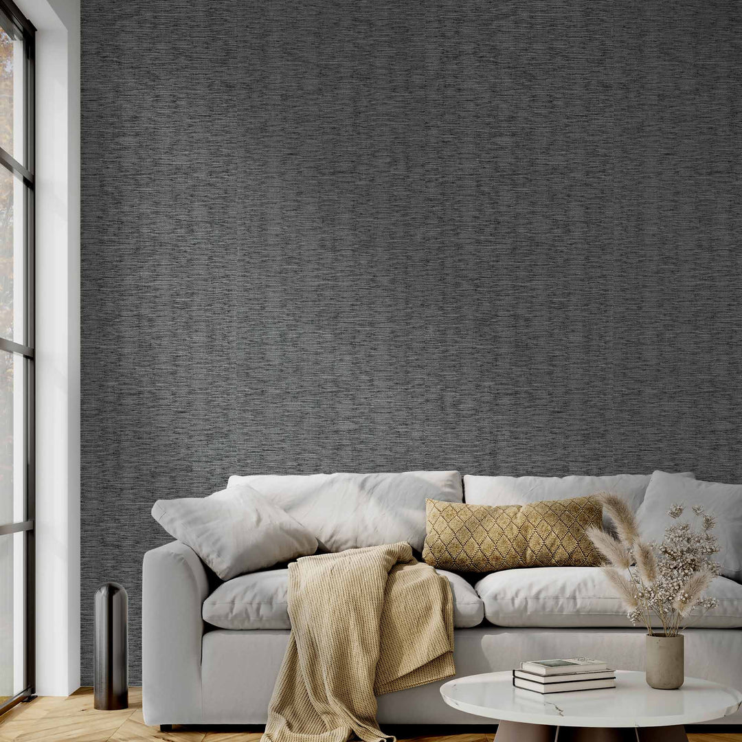 Modern living room with a light gray sofa, neutral cushions, and a beige throw blanket, set against a dark textured wallpaper. A round white coffee table with books and a vase of dried flowers is in front of the sofa, and large floor-to-ceiling windows on the left let in natural light.