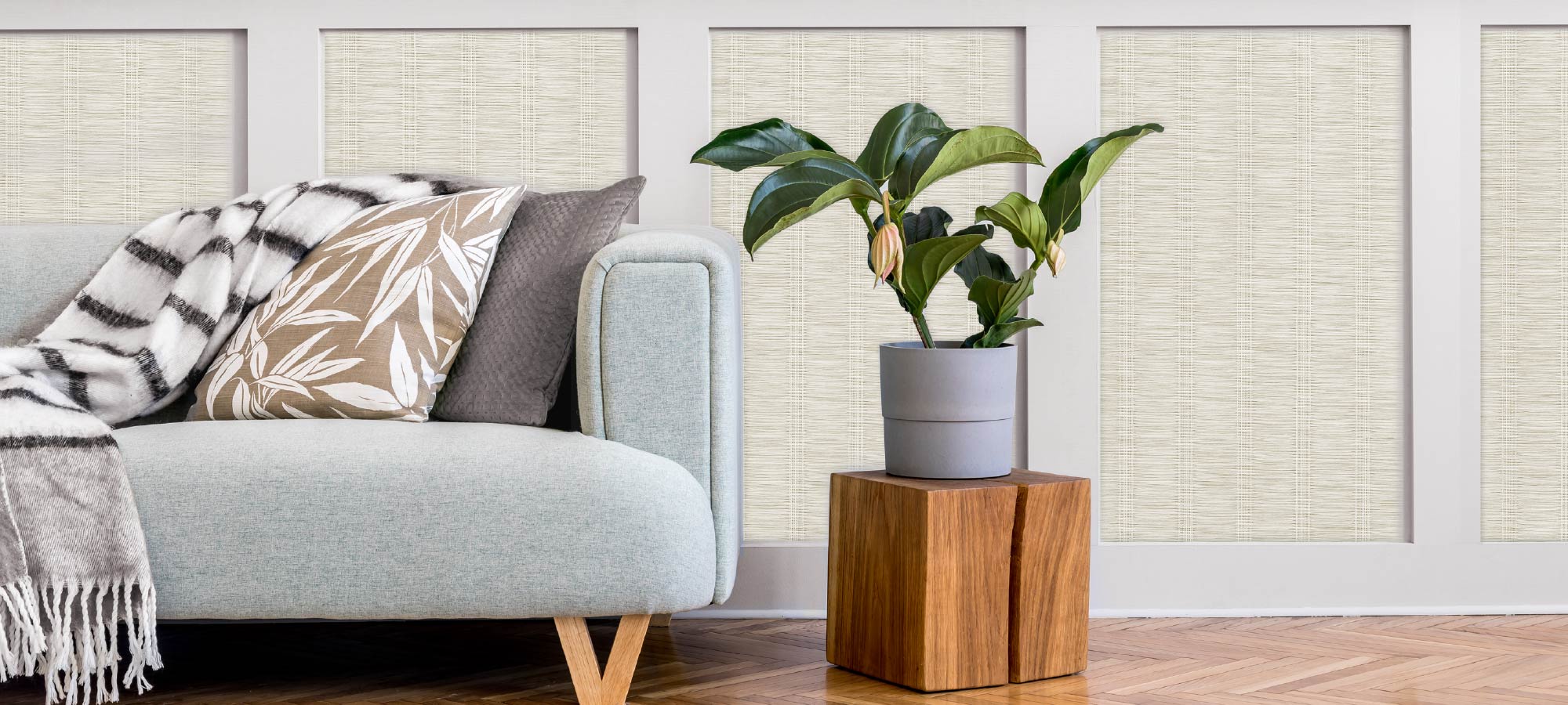 Cozy living room corner with light grey sofa, patterned cushions, striped throw blanket, indoor plant on wooden side table, and textured wall paneling.