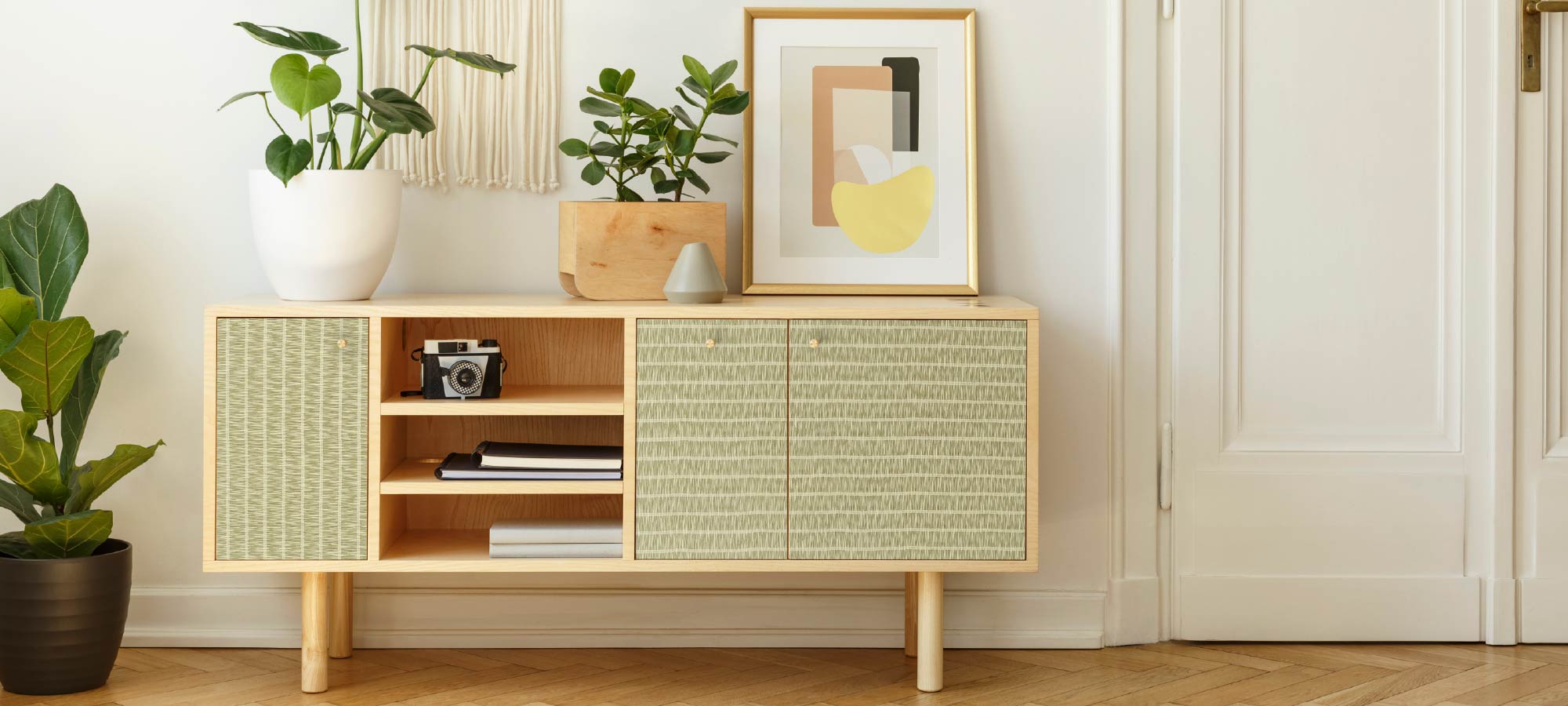Scandinavian-style sideboard with light green textured wallpaper panels, decorated with potted plants, framed abstract art, and wooden flooring.