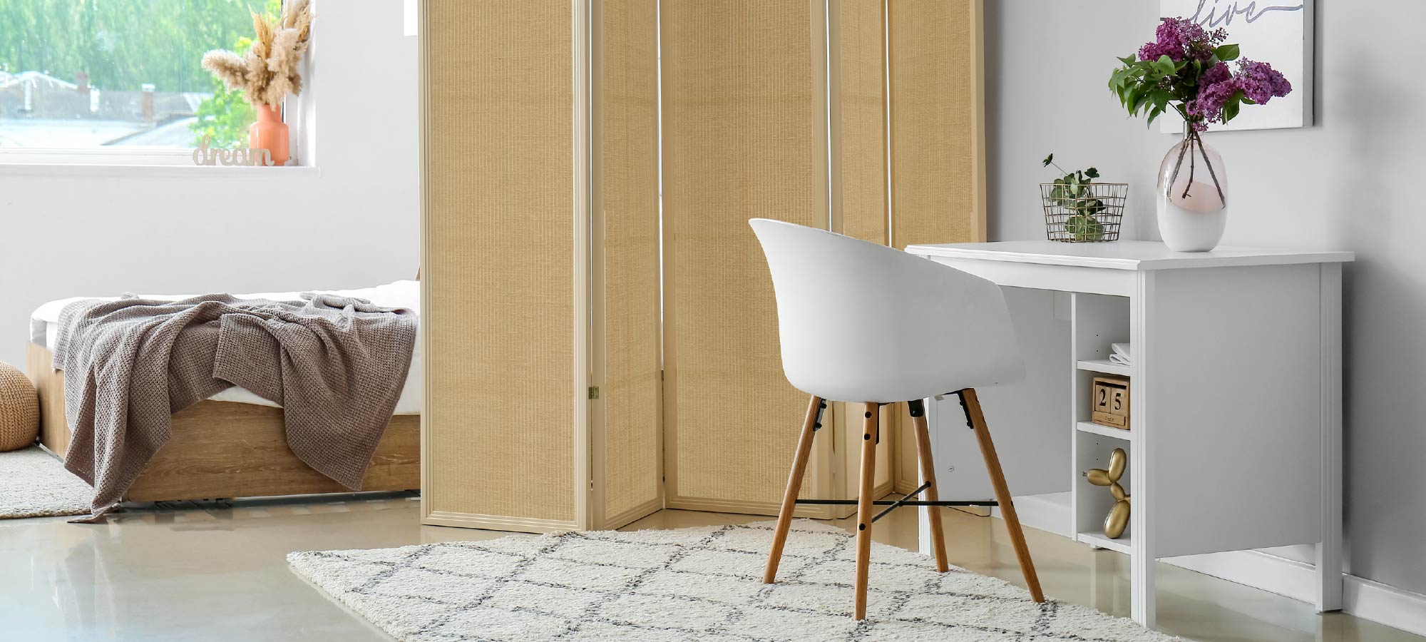 Cozy bedroom with natural woven textured wall divider, modern white desk and chair, soft bedding, decorative plants, and bright natural light from window.