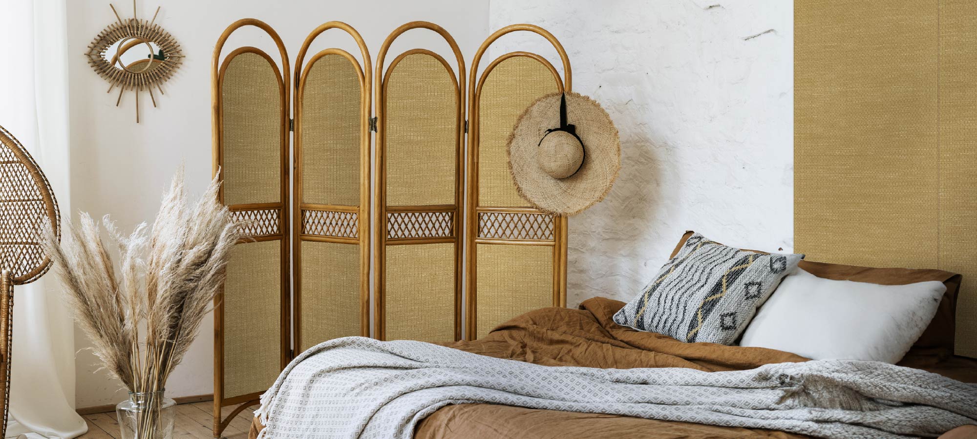 Bohemian style bedroom with rattan folding screen, woven wall accents, cozy bed with patterned pillows, and dried pampas grass in vase.