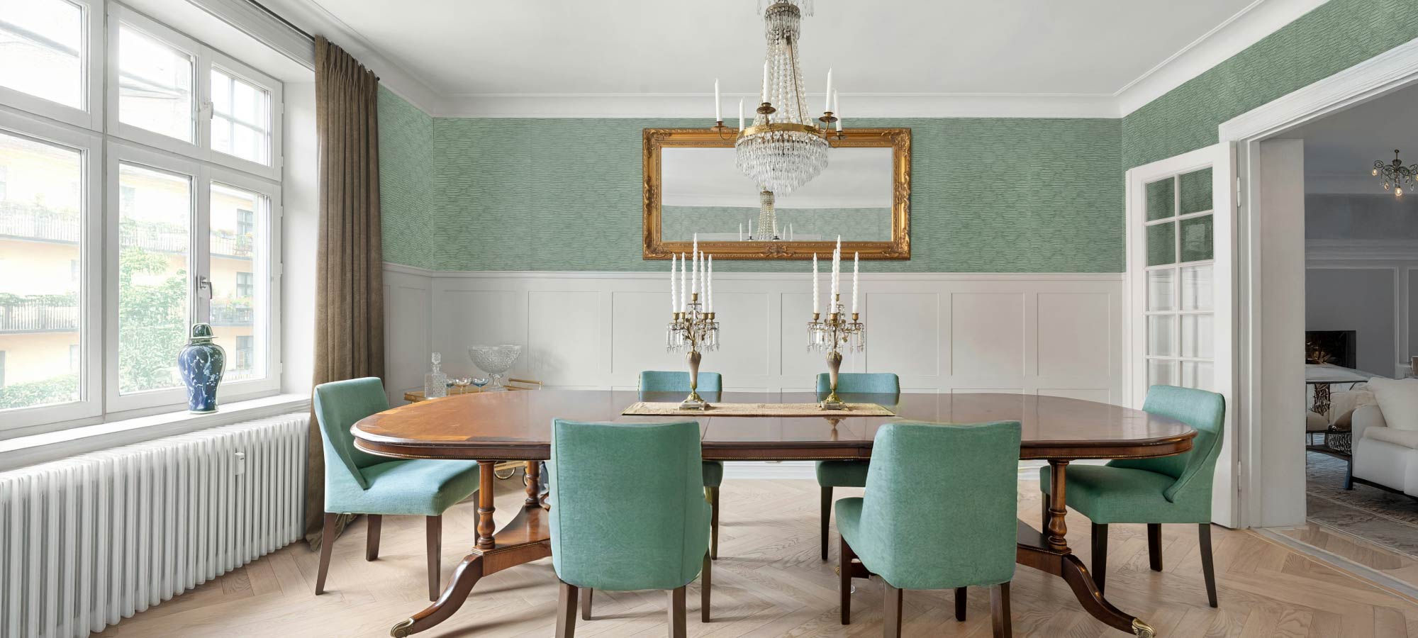 Elegant dining room with a classic wooden table, crystal chandelier, and upholstered chairs, featuring textured green accent walls and large windows for natural light.