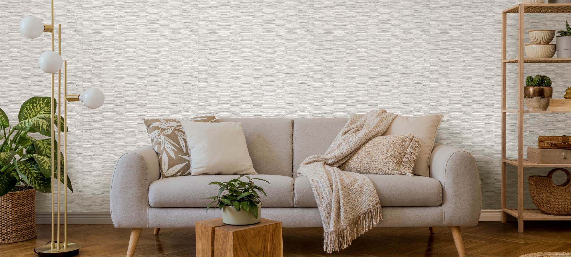 Cozy living room interior with beige sofa, textured throw blanket, patterned cushions, indoor plants, wooden side table, gold floor lamp, and light textured wall.