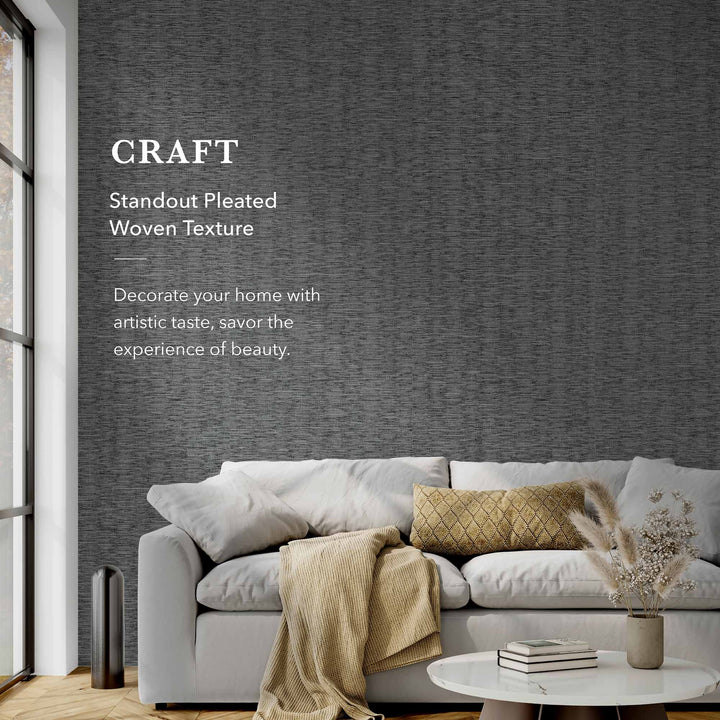 Modern living room featuring a light gray sofa with neutral cushions and a beige throw blanket, set against a dark gray pleated woven textured wallpaper. The text on the wall reads “CRAFT – Standout Pleated Woven Texture. Decorate your home with artistic taste, savor the experience of beauty.” A round white coffee table with books and a vase of dried flowers sits in front, and large floor-to-ceiling windows on the left bring in natural light.