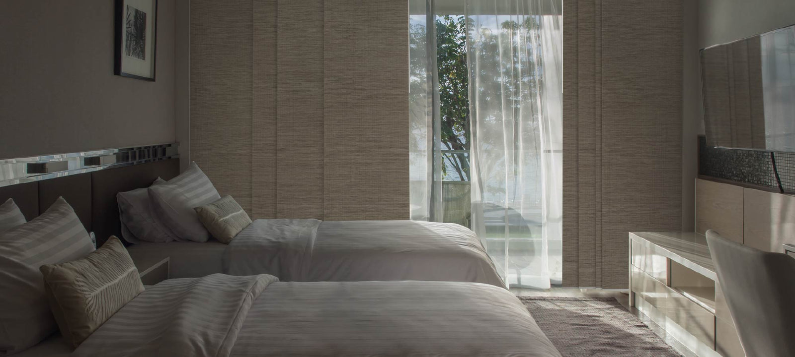 Modern bedroom with two twin beds facing a large sliding glass door covered by textured beige panel track blinds and sheer white curtains, letting in natural light and a view of trees outside.