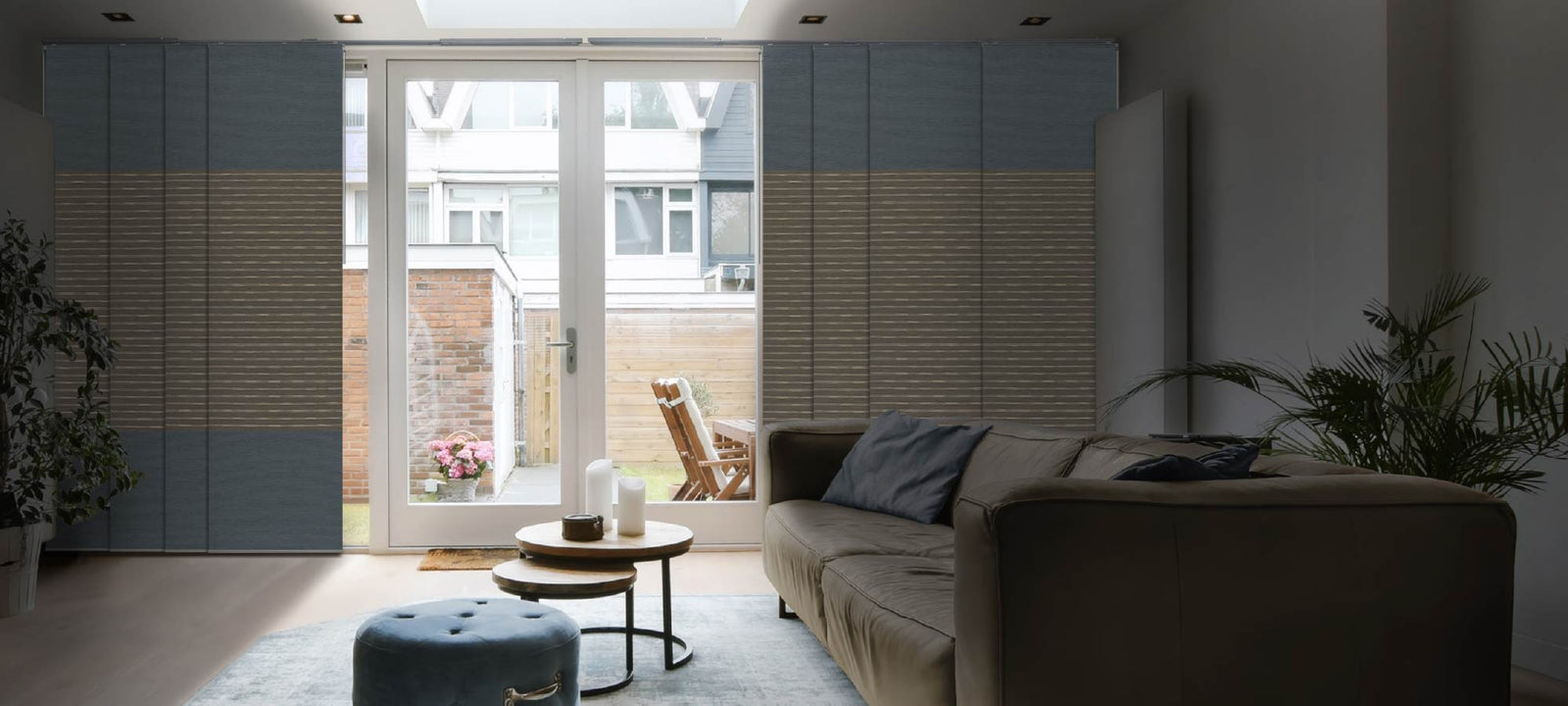Contemporary living room with full-height layered fabric panels in two-tone blue and taupe, filtering light beside a wide glass door; space features a plush taupe sofa, round nesting coffee tables, a velvet ottoman, and indoor greenery facing a bright patio.