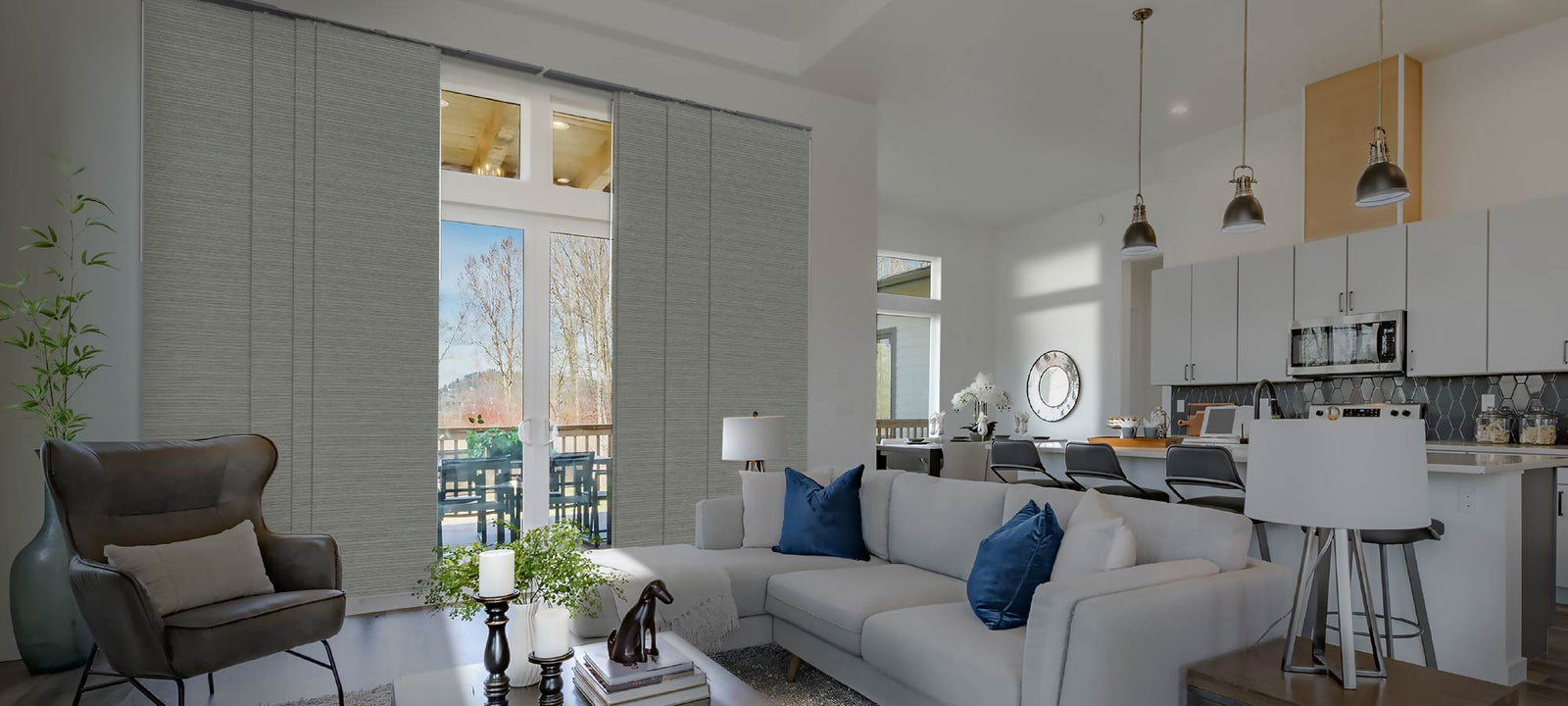 Bright and airy open-plan living space with soft textured panels covering tall glass doors; modern kitchen with white cabinetry, navy accents on a sectional sofa, and a cozy reading nook with a leather armchair and indoor greenery.