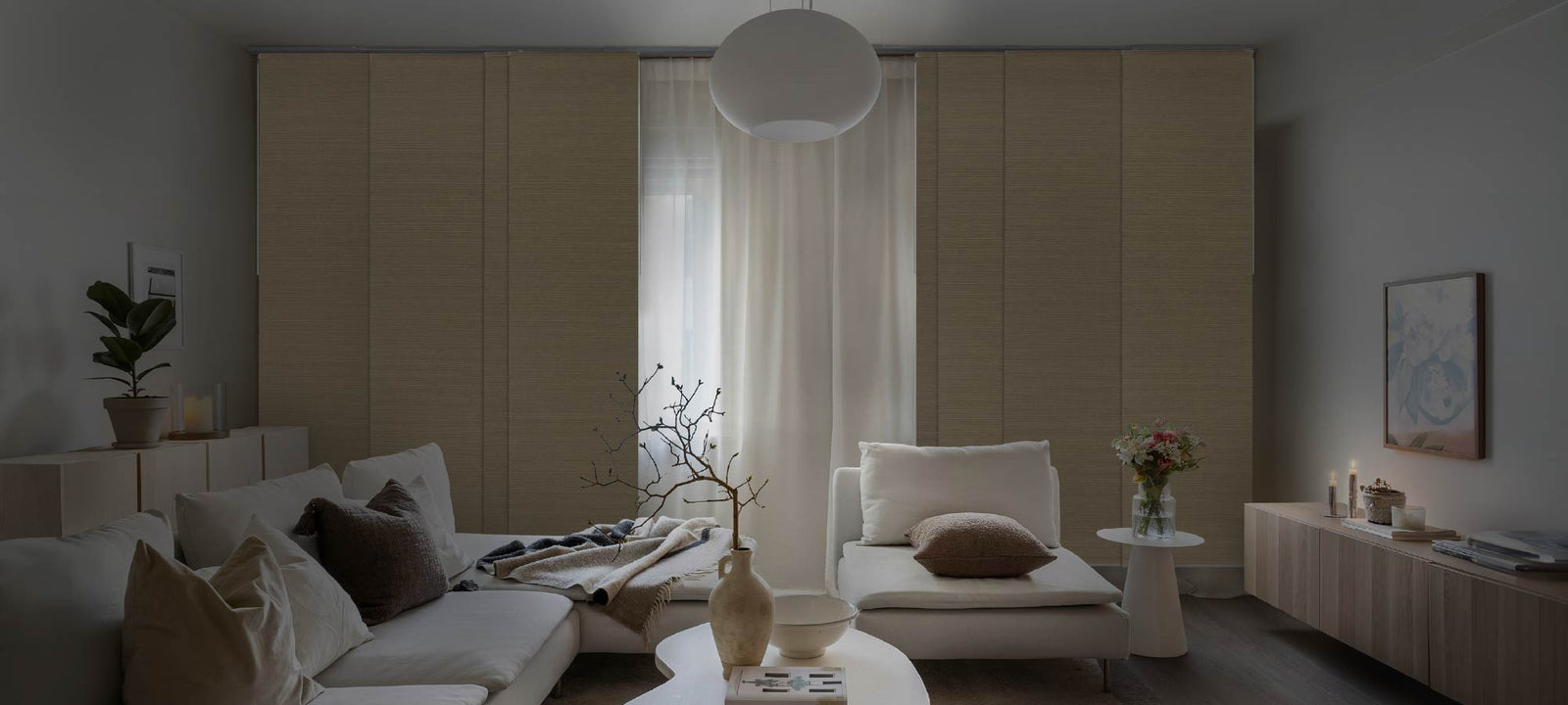 Cozy living room styled with layered neutral cushions, a ceramic vase with branches, soft overhead lighting, and floor-to-ceiling textured panels filtering natural light for a warm, tranquil atmosphere.
