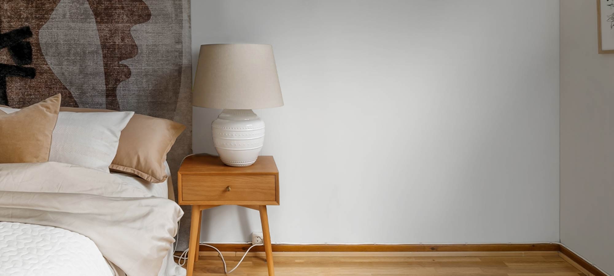 Minimalist bedroom with wooden nightstand, ceramic table lamp, neutral bedding, and abstract wall art for a cozy modern interior.