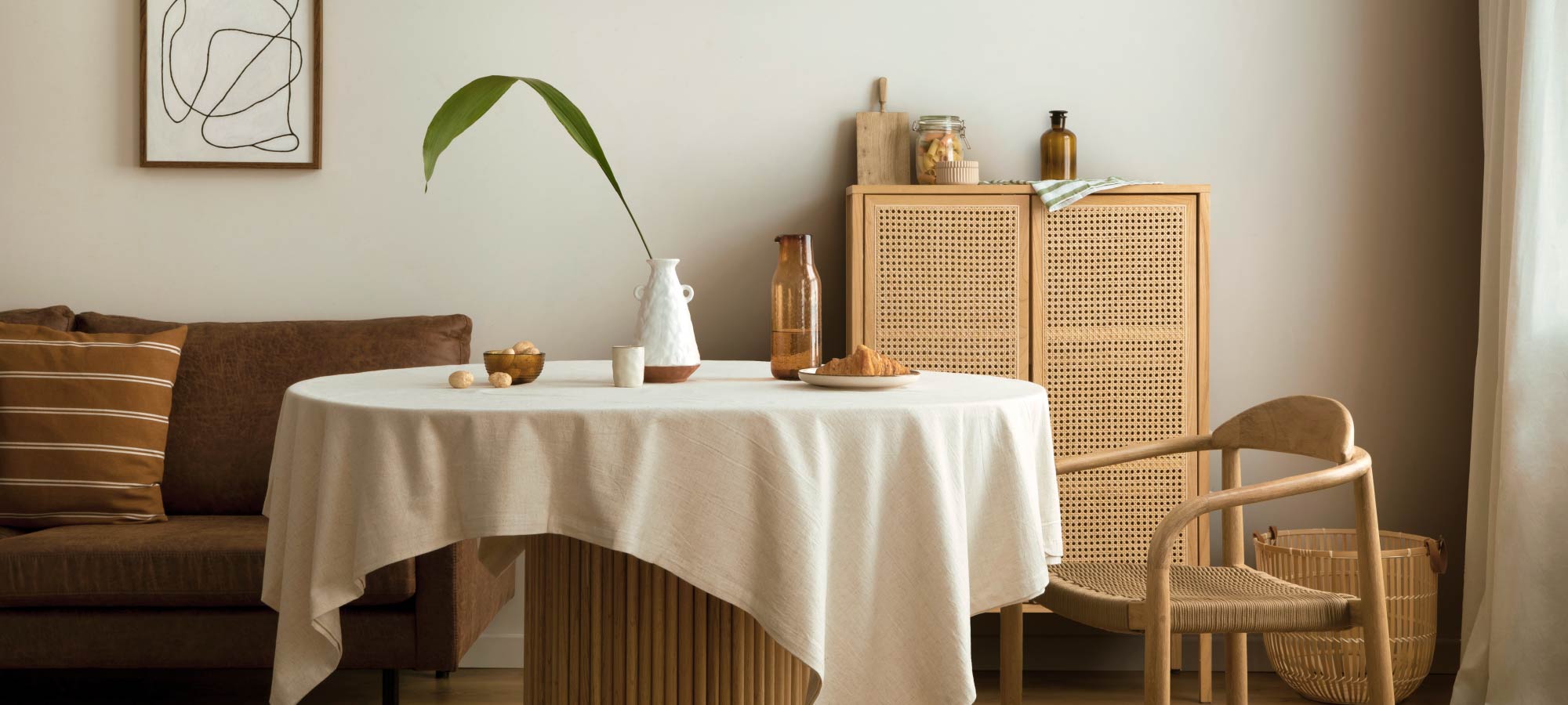 Cozy dining room with round table covered in linen tablecloth, rattan chair, wooden cabinet, brown sofa, and warm minimalist home decor.