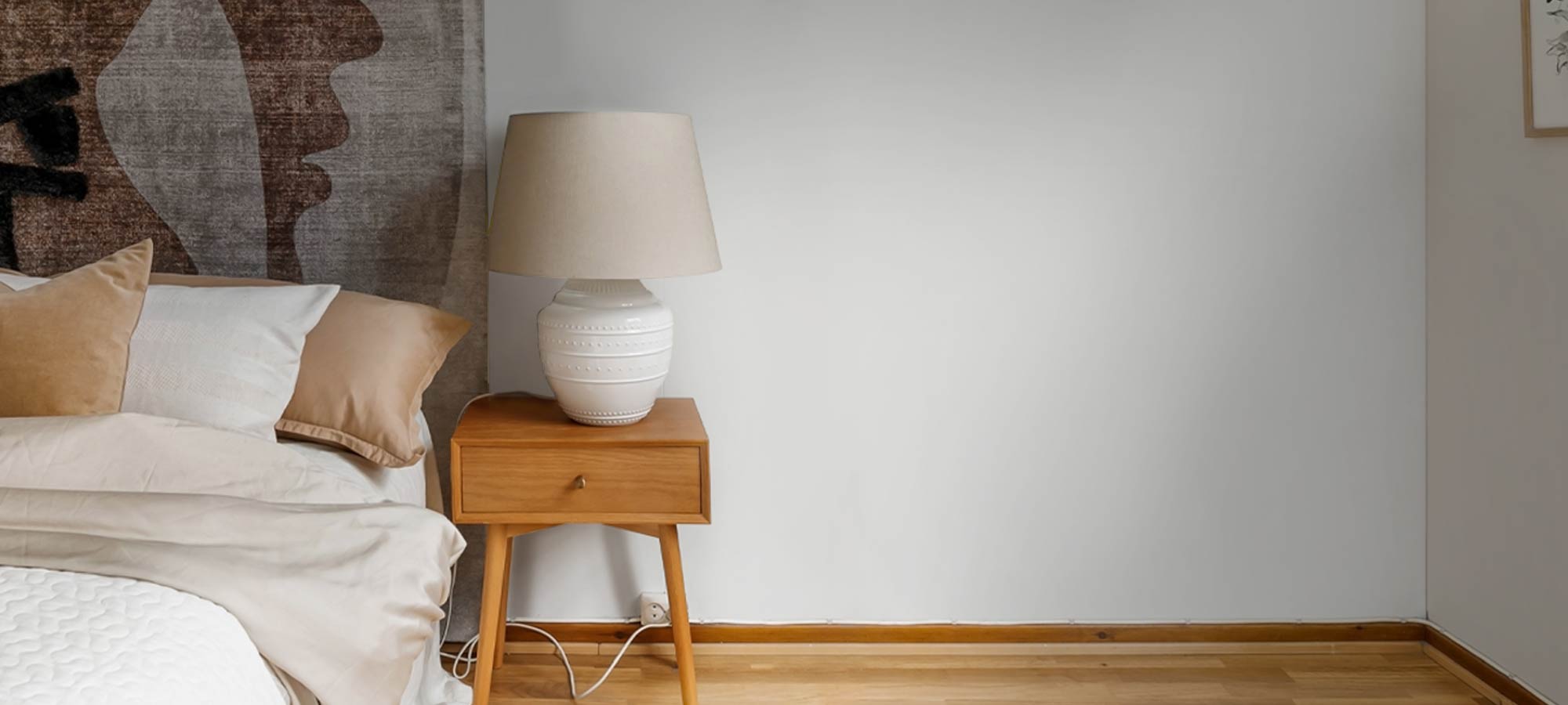 Bright bedroom interior with wooden flooring, clean white wall, and simple nightstand lamp setup.