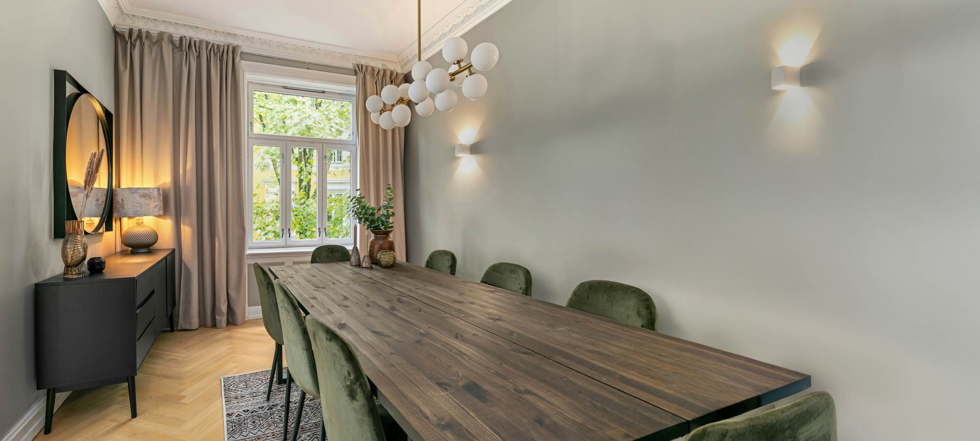 Modern dining room with a long dark wood table, velvet chairs, and soft green walls, styled with a statement chandelier, neutral curtains, and warm ambient lighting.