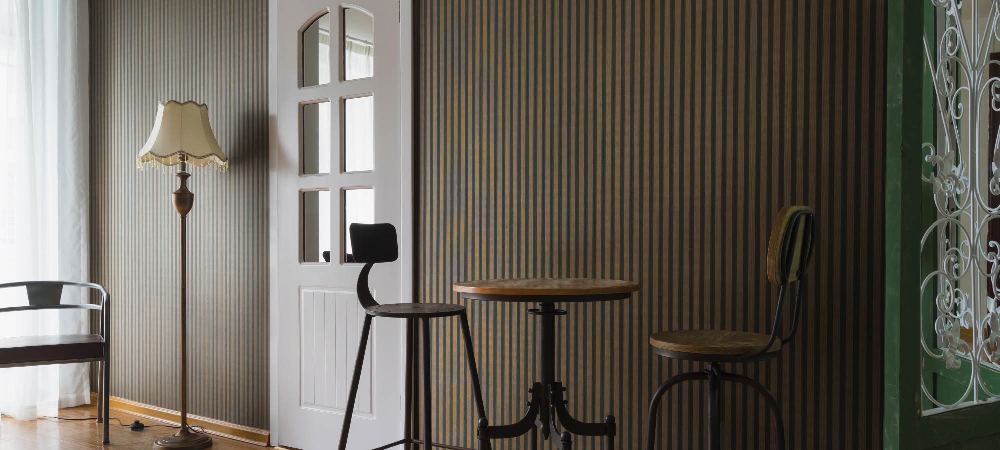 Vintage-inspired cafe corner with striped accent wall, tall bar stools, round wooden table, floor lamp, and white door with glass panels.