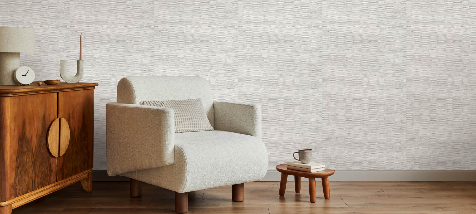 Modern living room with beige fabric armchair, small wooden coffee table with books and mug, vintage wooden cabinet with lamp and decor, and light textured wall background.