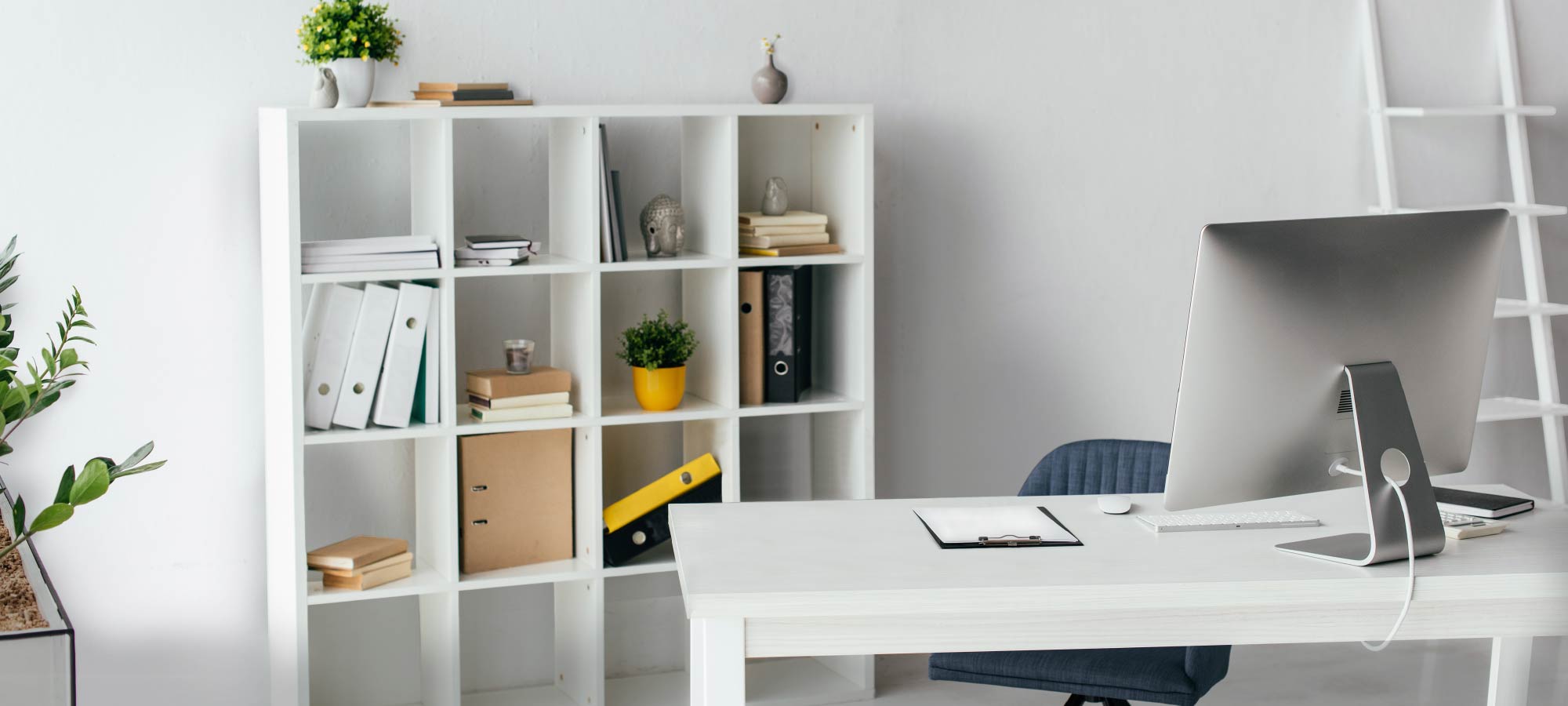 Minimalist home office interior with clean white desk, modern computer setup, organized cube bookshelf, green indoor plant, and neutral wall background.