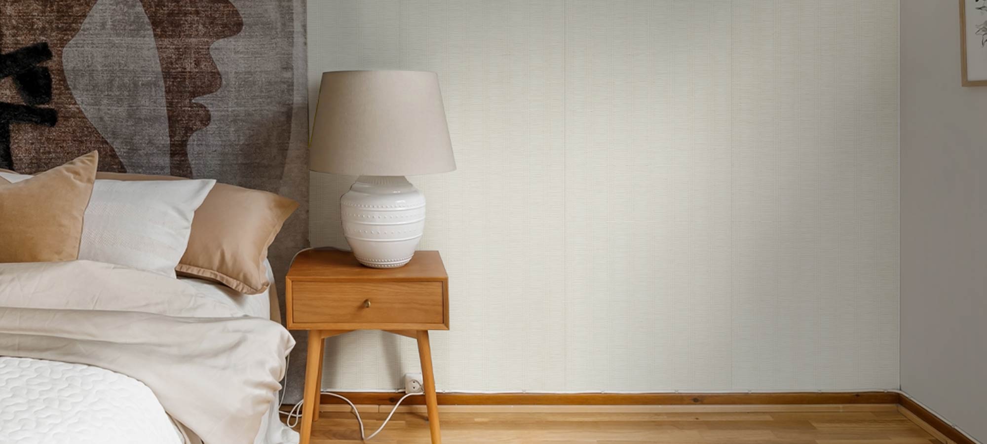 Modern bedroom with wooden nightstand, ceramic table lamp, neutral bedding, and light textured wallpaper for a cozy minimalist interior.