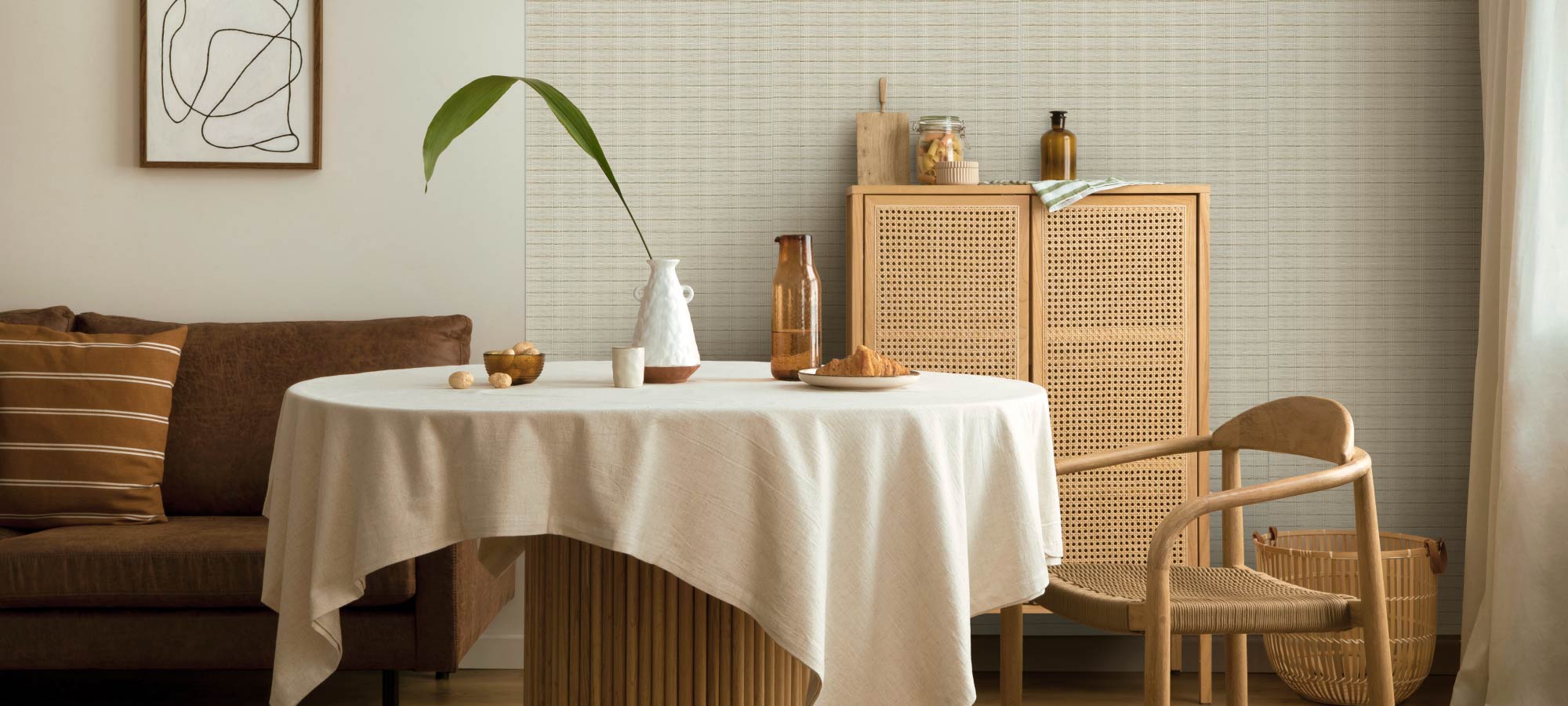 Stylish dining area with linen-covered round table, rattan chair, woven cabinet, brown sofa, and textured accent wall creating a cozy modern home atmosphere.