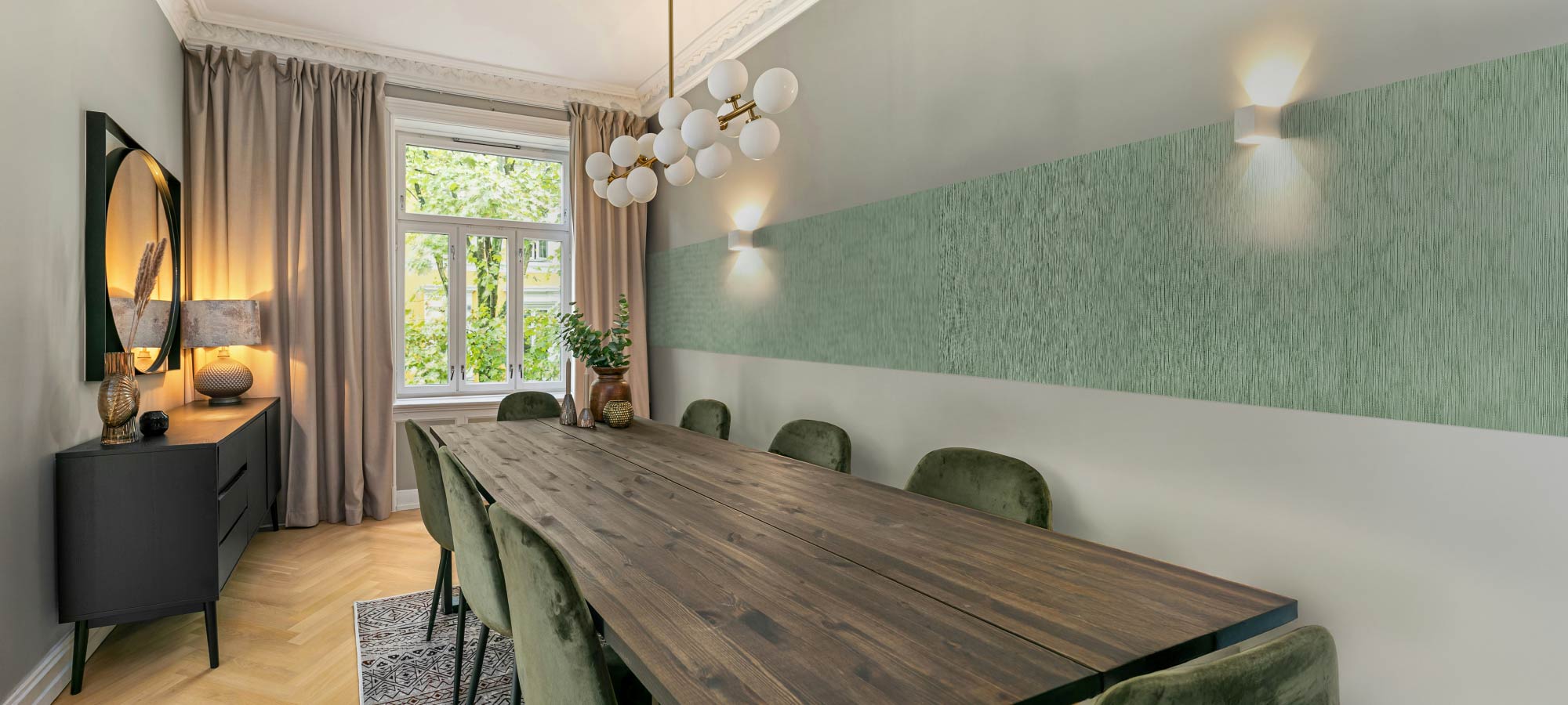 Elegant dining space with a long wooden table, velvet chairs, and a textured green accent wall highlighted by warm wall lights and natural window light.