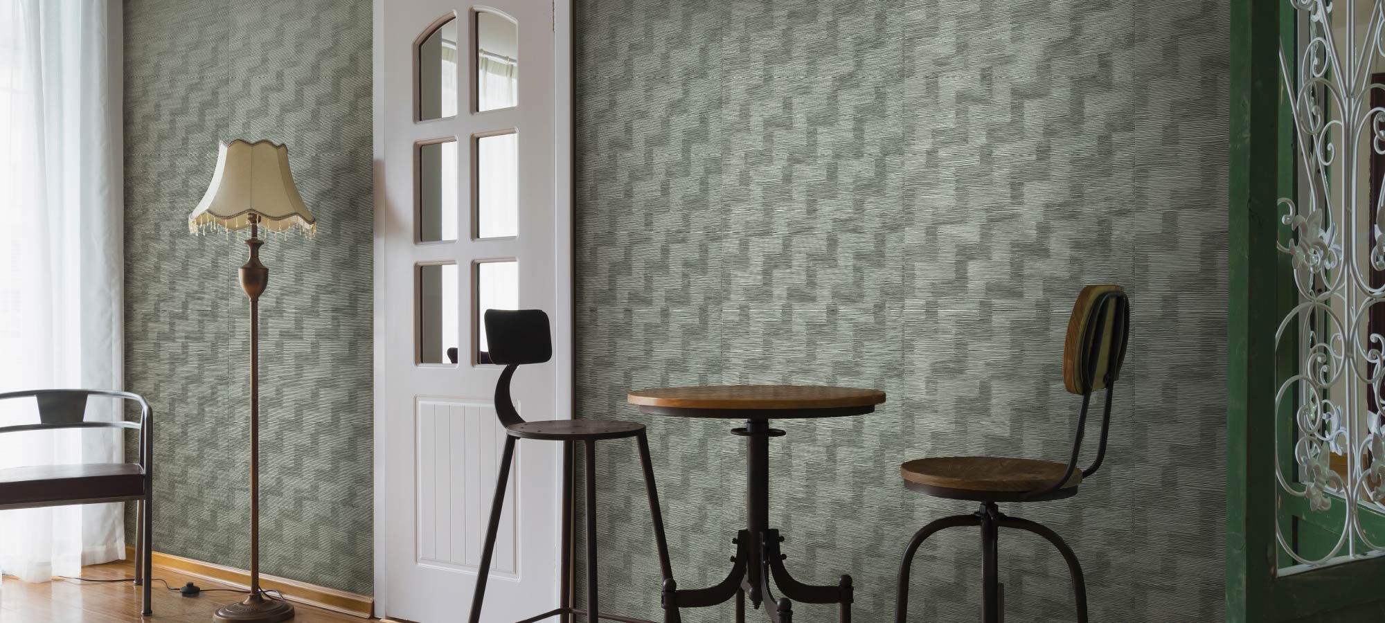 Cozy corner interior with patterned green accent wall, round wooden bar table, vintage-style stools, white door with glass panels, and classic floor lamp.