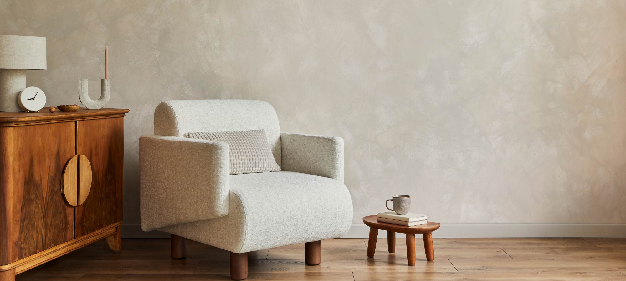 Minimalist living room with beige textured wall, modern fabric armchair with cushion, small wooden side table with books and ceramic mug, and vintage wooden cabinet with table lamp decor.