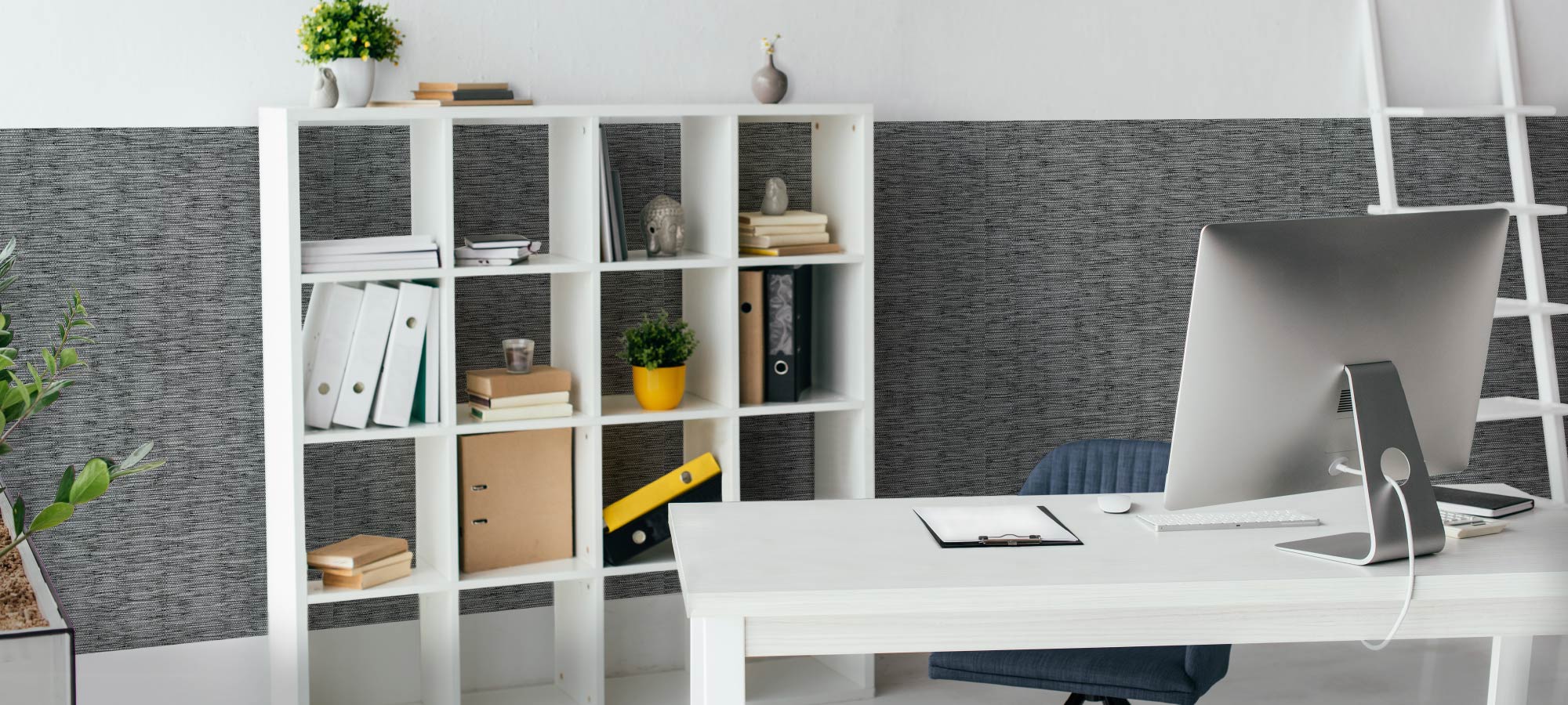 Contemporary office space featuring white desk with computer, organized cube bookshelf with folders and plants, stylish dark textured accent wall, and minimalist interior design.