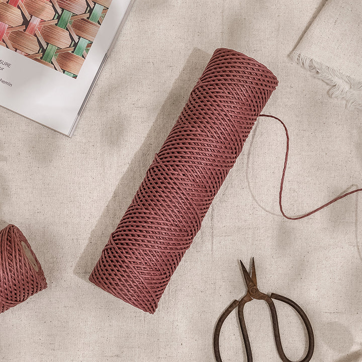 Godear Design_A roll of red grapefruit paper raffia ribbon next to a pair of scissors on a beige background, with a part of the ribbon unrolled to show the texture.